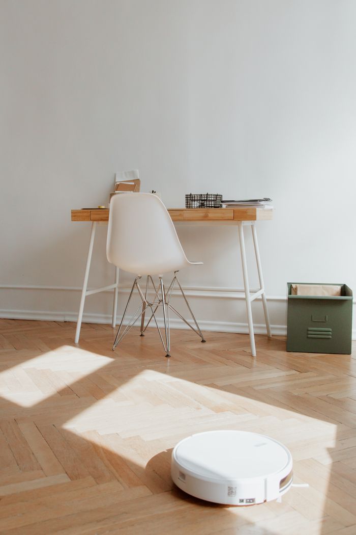 A modern home office setup featuring a desk and robotic vacuum on a wooden floor, highlighting minimalism.